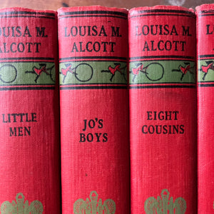 antique book set in red and green - Classic American Literature Book Set Written by Louisa May Alcott - Little Women, Little Men, Jo's Boys, Eight Cousins, Rose in Bloom and An Old-Fashioned Girl - cover art by Anna Bond - closeup view of the spines showing an image of children playing with a hoop & stick