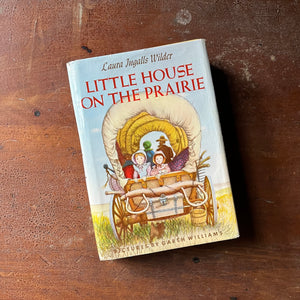 vintage children's chapter book - Little House on the Prairie written by Laura Ingalls Wilder with illustrations by Garth Williams - view of the dust jacket's front cover with an illustration of Mary & Laura in the back of a covered wagon with Jack the Bulldog under the wagon - Ma & Pa can be seen in the foreground
