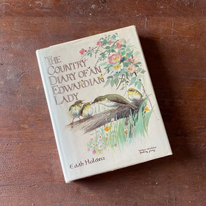 vintage nature sketchbook & diary - The Country Diary of An Edwardian Lady written & illustrated by Edith Holden - view of the dust jacket's front cover with an illustration of a momma bird feeding two baby birds surrounded by flowers & grass