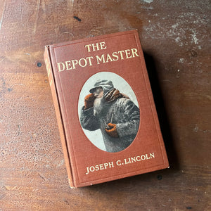 antiquarian book, novel - The Depot Master written by Joseph C. Lincoln with illustrations by Howard Heath - view of the front cover with an illustration of the Depot Master on it - he is holding a pipe & wearing a hat, jacket, mittens, scarf & glasses