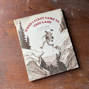 vintage children's picture book, folk song - When I First Came to this Land written by Oscar Brand with illustrations by Doris Burn - view of the dust jacket's front cover with a man stepping over a canyon with everything he owns on his back